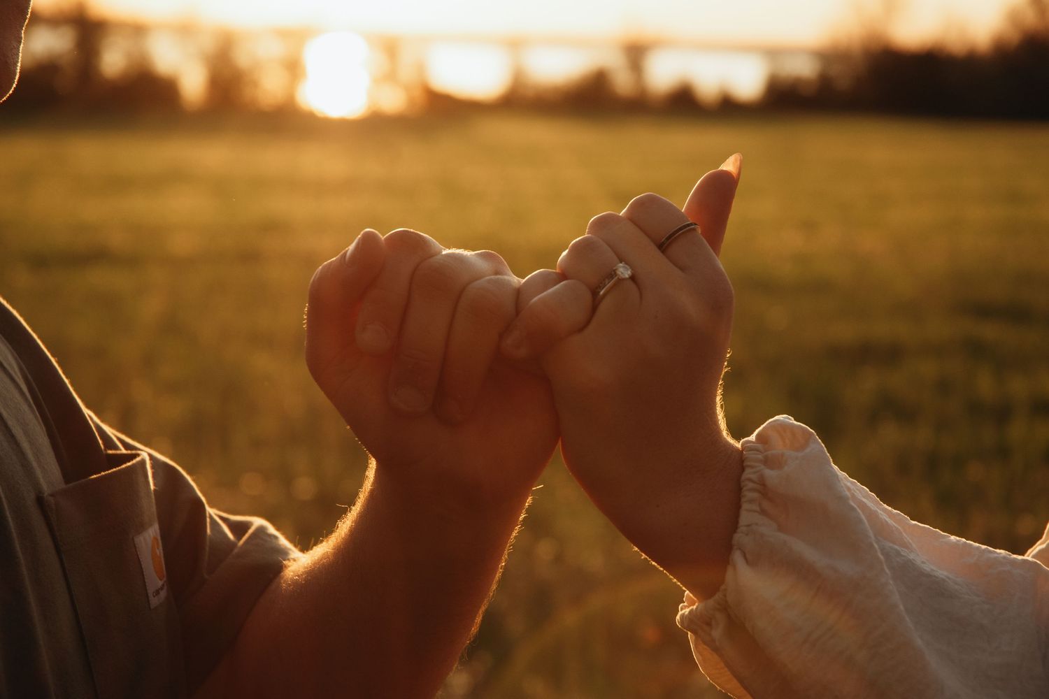 Couple holding hands with engagement ring at sunset in a golden field.