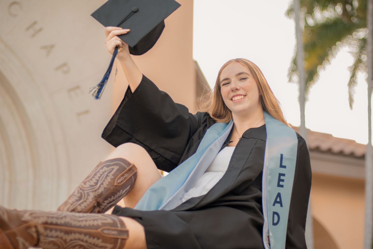 Smiling female graduate in black cap and gown holding mortarboard aloft outdoors near chapel with LEAD sash.
