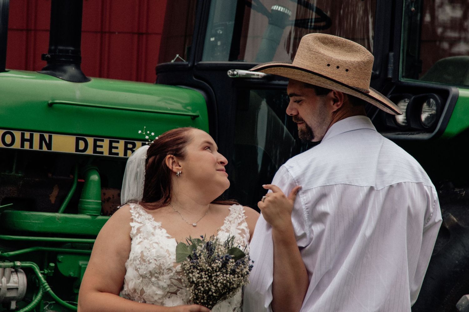Romantic couple in wedding attire standing near a John Deere tractor in a rustic outdoor setting.