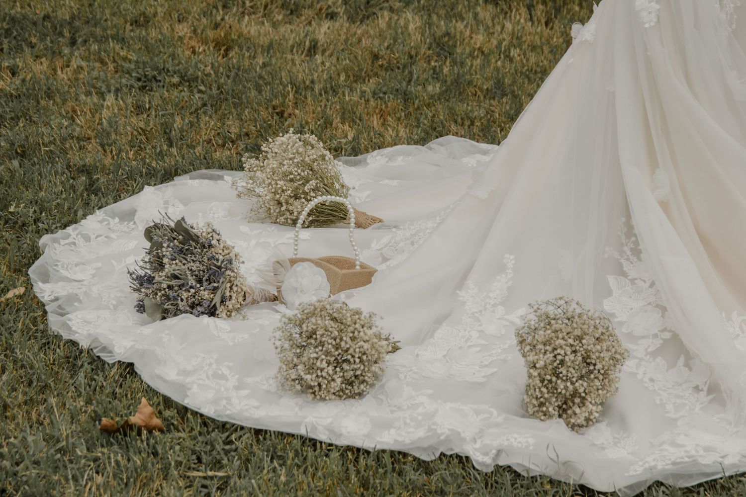 A delicate white wedding dress train with baby's breath flowers lies gracefully on green grass in natural lighting.