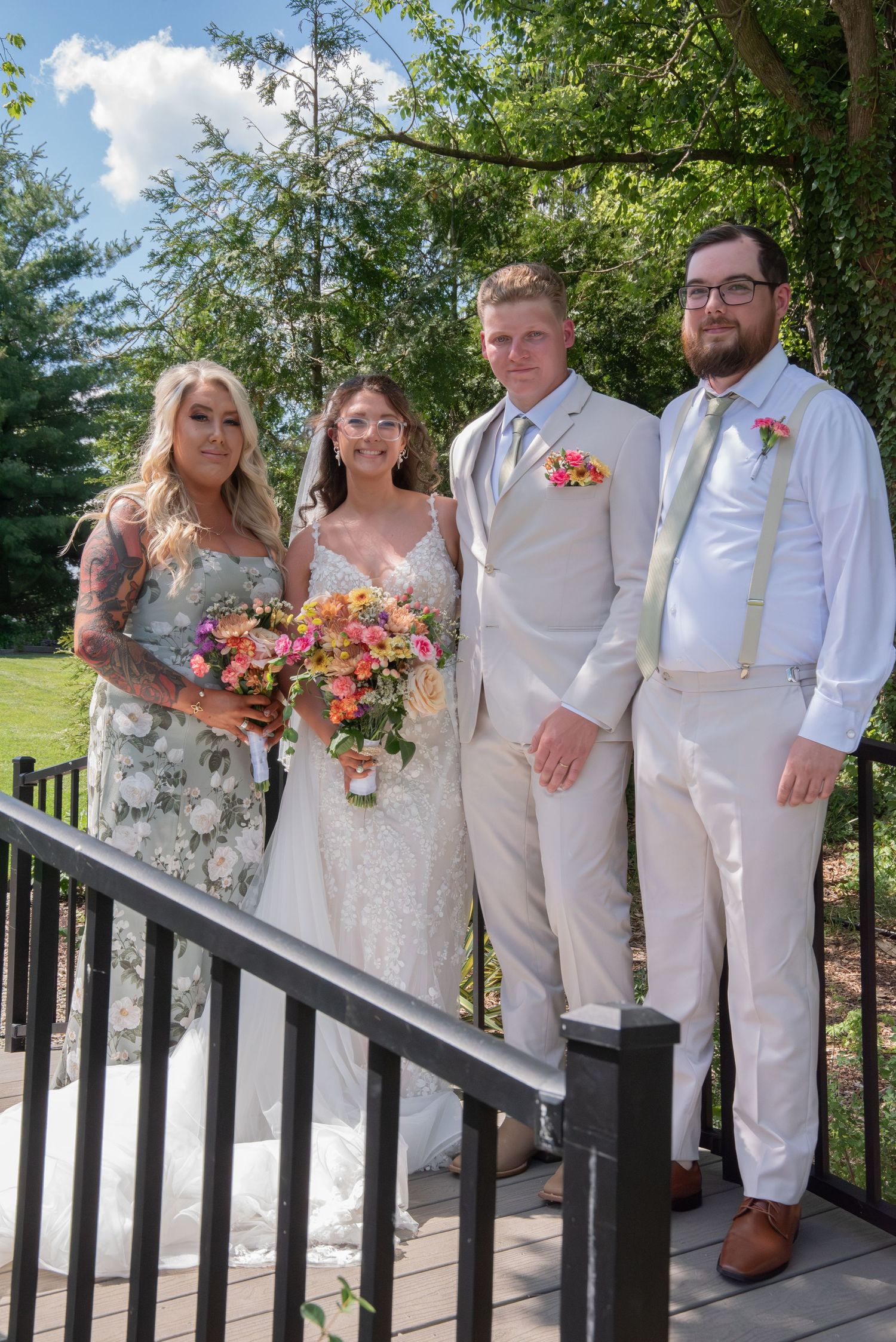 Wedding party of four posing on a deck, bride in white lace gown with bouquet, groom in tan suit with floral boutonniere.