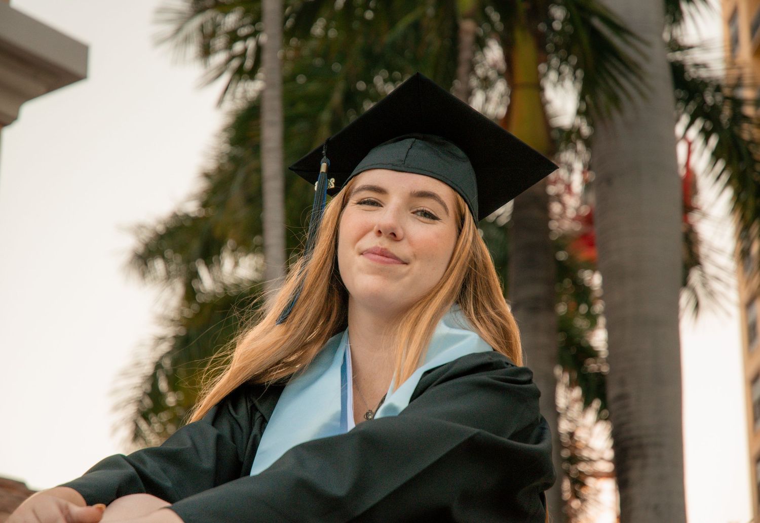 Smiling female graduate in black cap and gown with light blue tassel poses outdoors near palm trees.