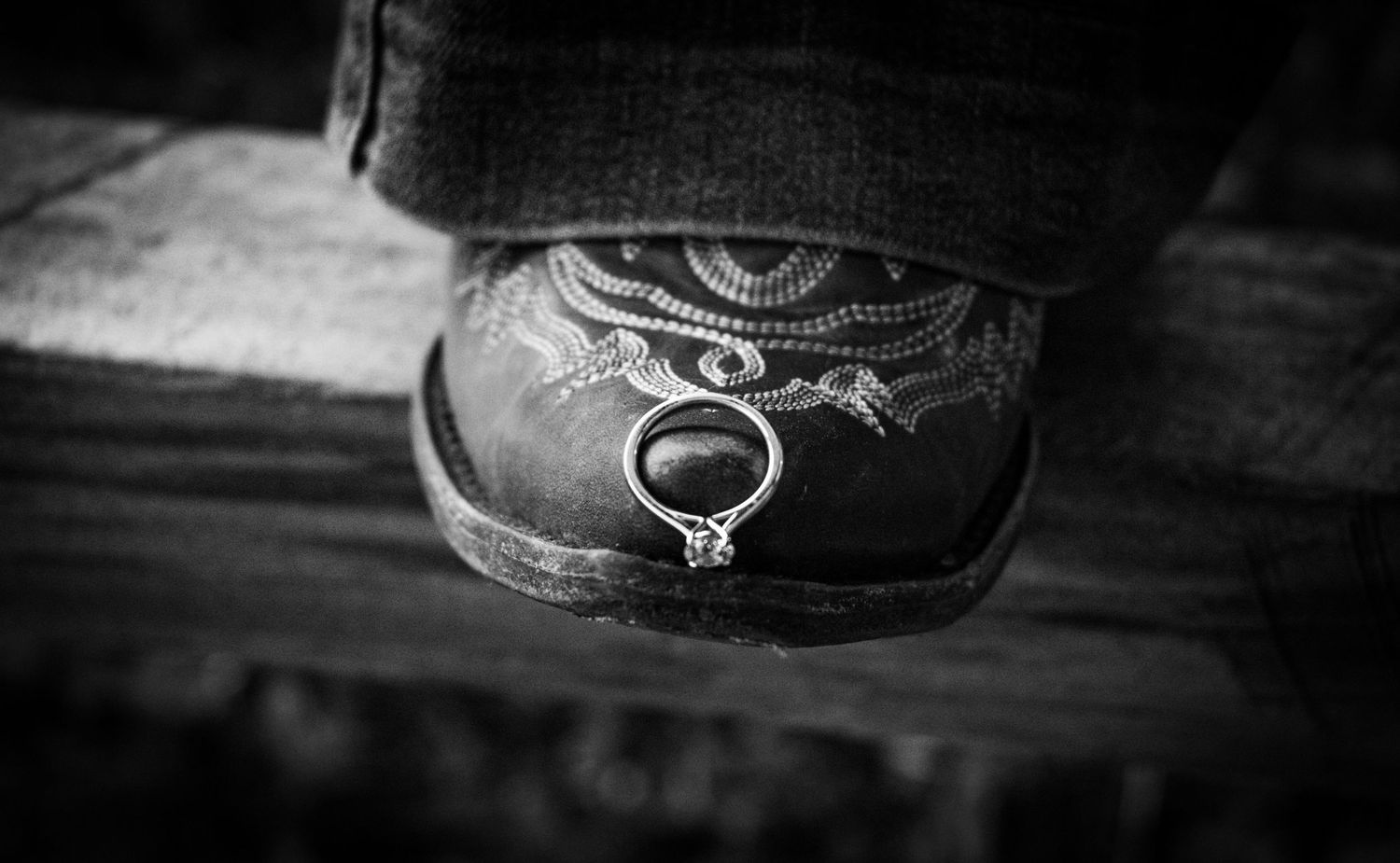 Black and white photo of a diamond engagement ring resting on the toe of a decorative cowboy boot.