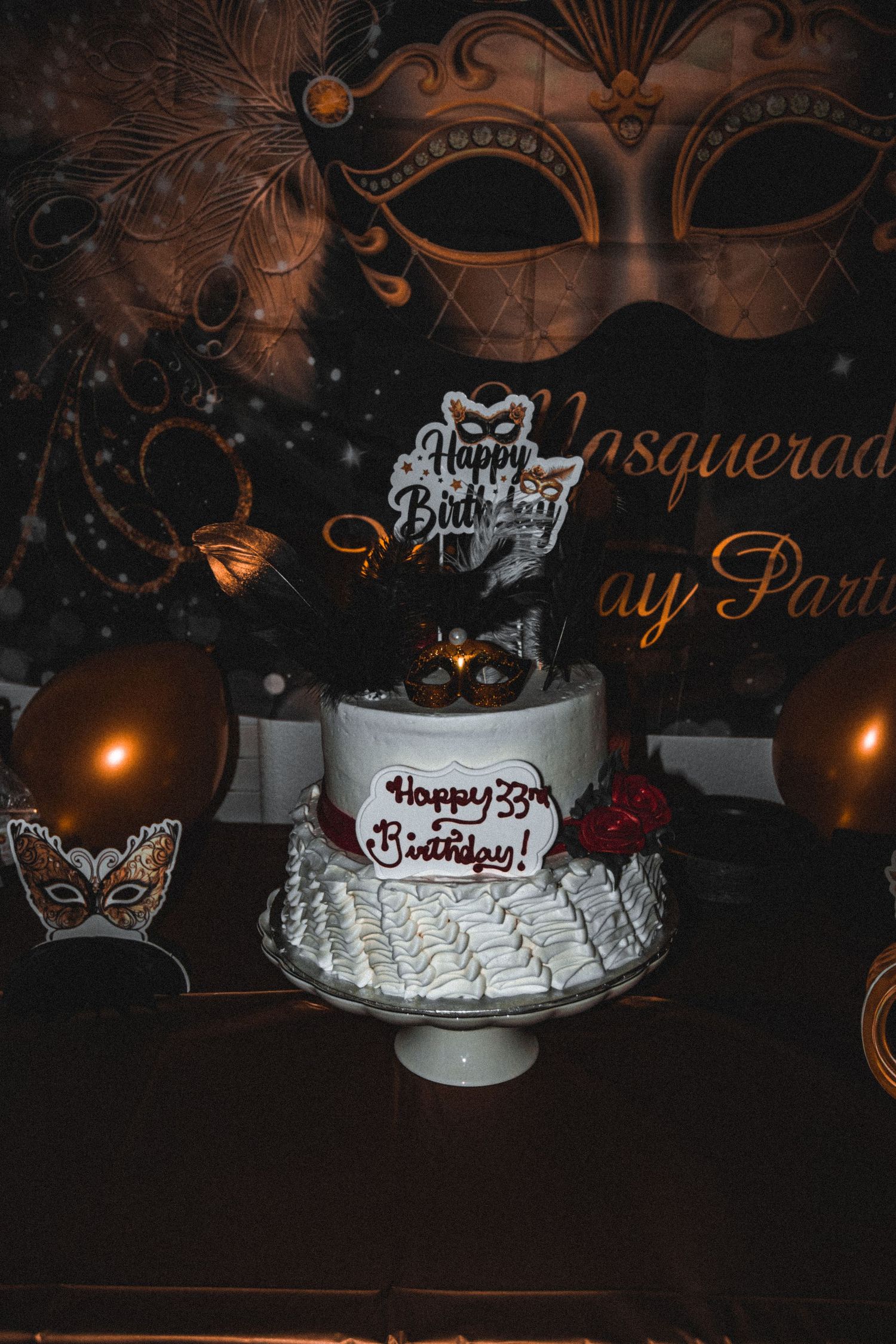 A white birthday cake with masquerade mask decorations and candles on a dark themed party table.