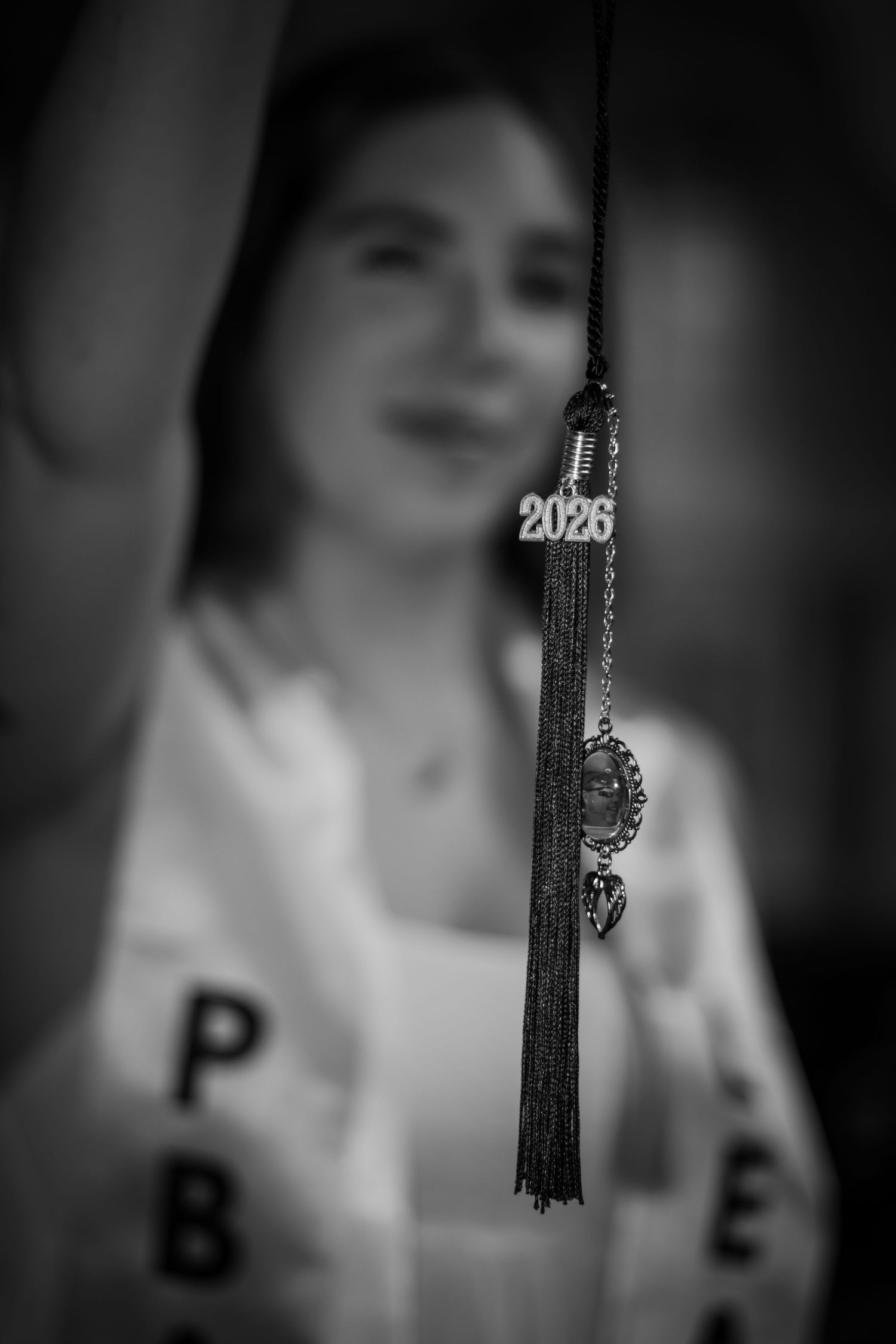 Black and white photo of a 2026 graduation tassel with charms in focus, smiling graduate blurred in background.