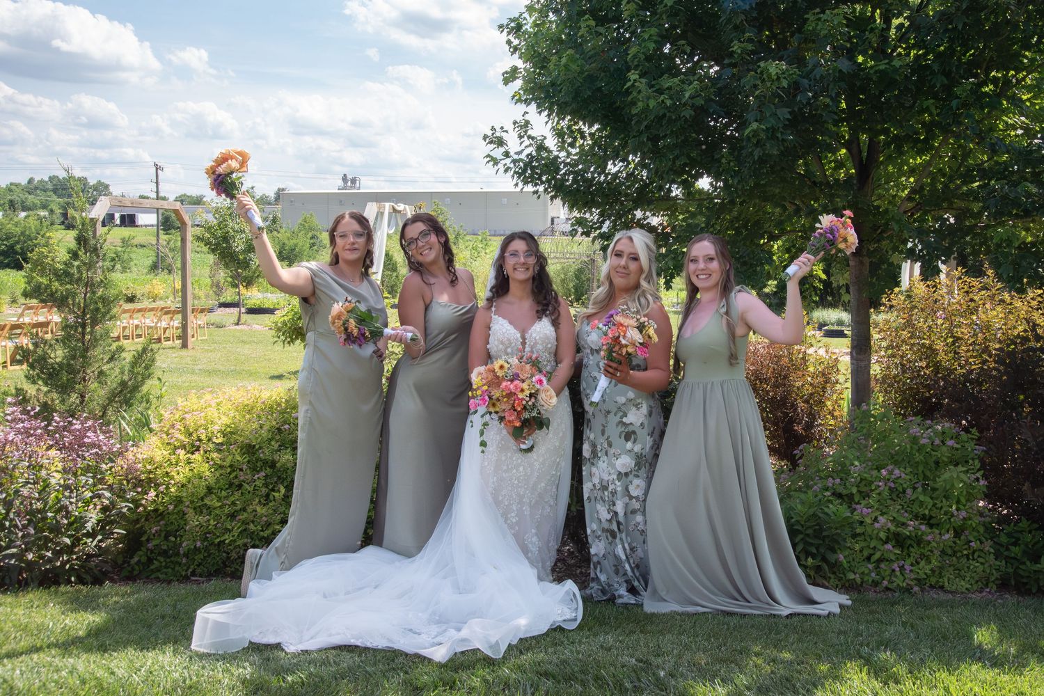 Bride in white gown poses with four bridesmaids in sage green dresses holding colorful bouquets outdoors.