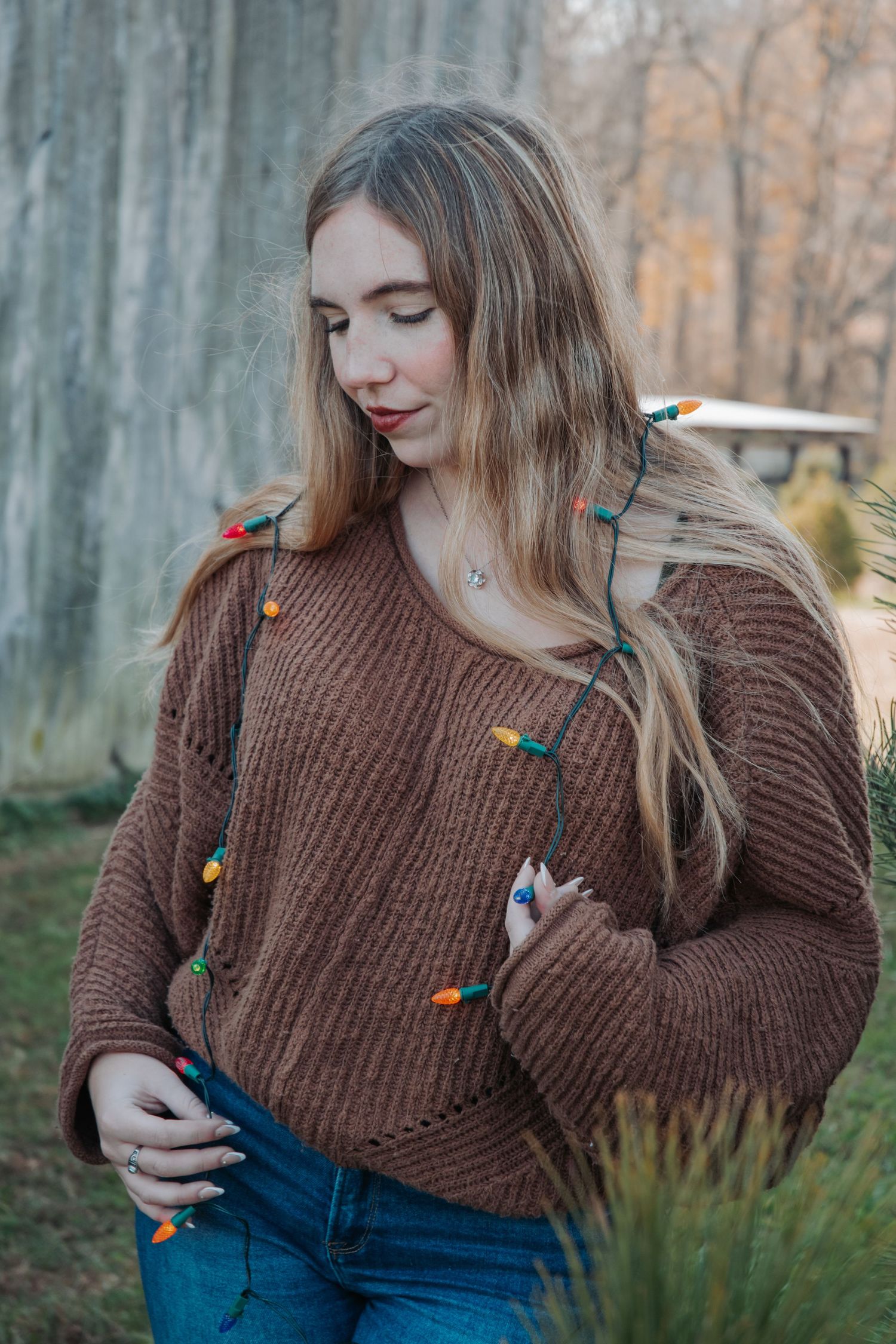 Woman in brown sweater with holiday lights wrapped around her during winter photoshoot.