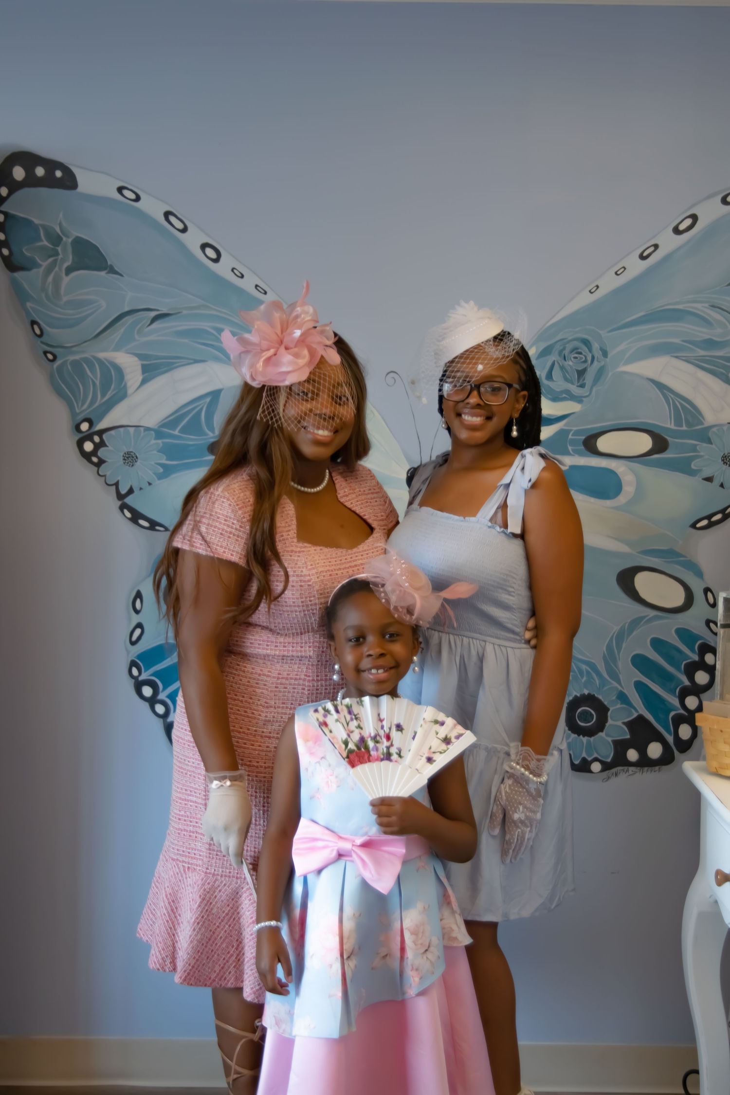 Three guests pose in front of large blue butterfly wings at a tea party celebration.