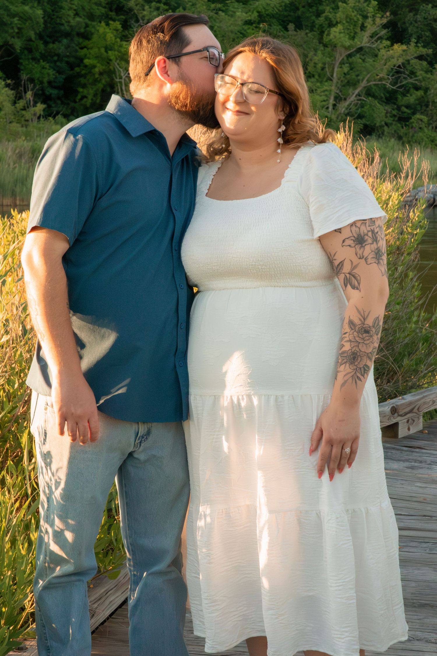 A couple poses outdoors on a wooden bridge, man kissing woman's cheek, she wears a white dress with floral arm tattoo.