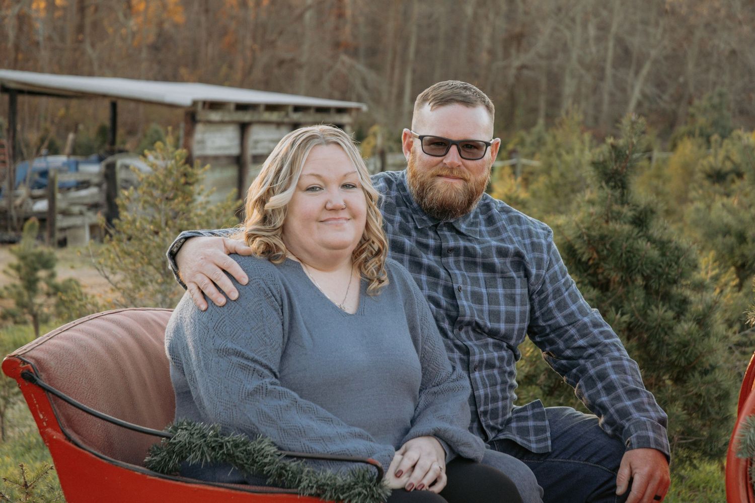 Couple poses together in an autumn outdoor setting with evergreen trees in the background.