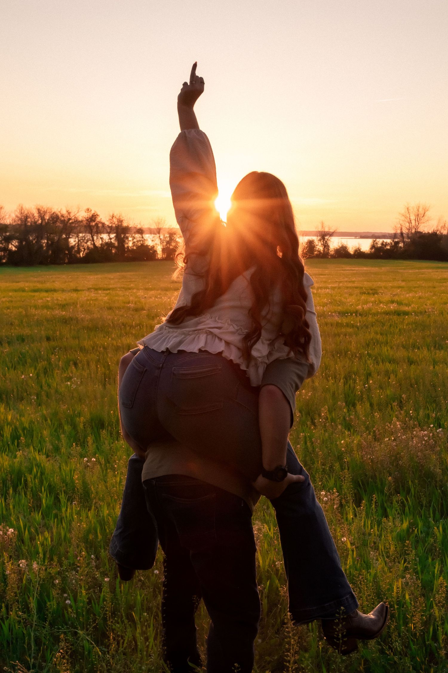 Couple sharing a romantic piggyback ride in a green field at golden sunset, woman pointing toward the sky.