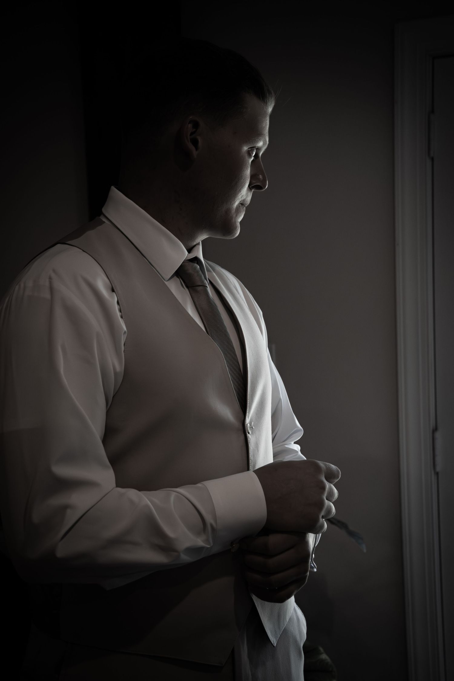 A groom in a white shirt, vest, and tie adjusts his cufflinks in dramatic low-key lighting near a window.