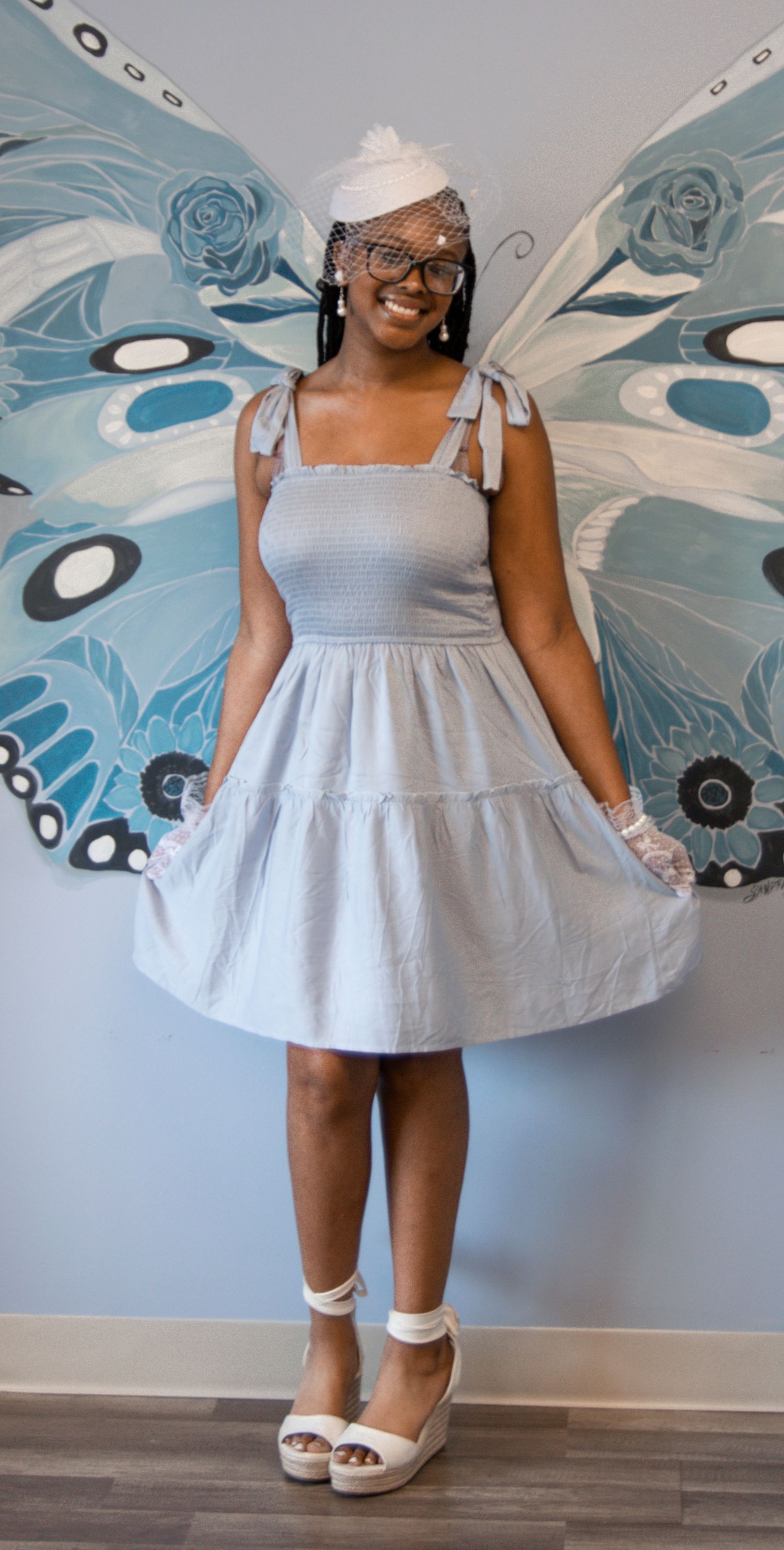 A guest in a light gray dress poses in front of large decorative butterfly wings.