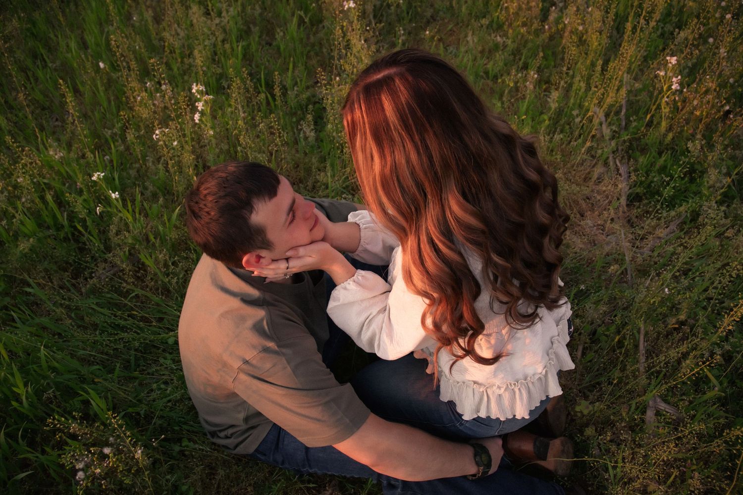 Romantic couple sitting in green meadow, woman with long red hair tenderly touching man's face at golden hour.