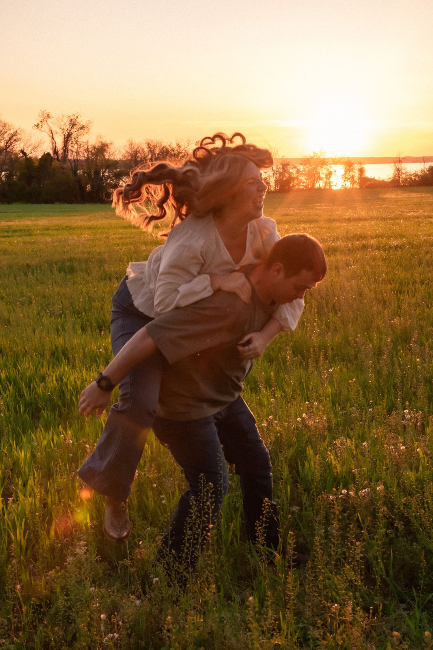 A couple joyfully playing piggyback in a green field during a golden sunset, with the woman's hair flowing in the wind.
