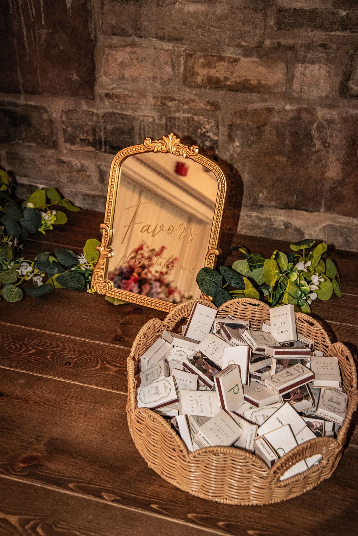 Wicker basket filled with wedding favor matchboxes beside a gold ornate mirror with Favors written on it.