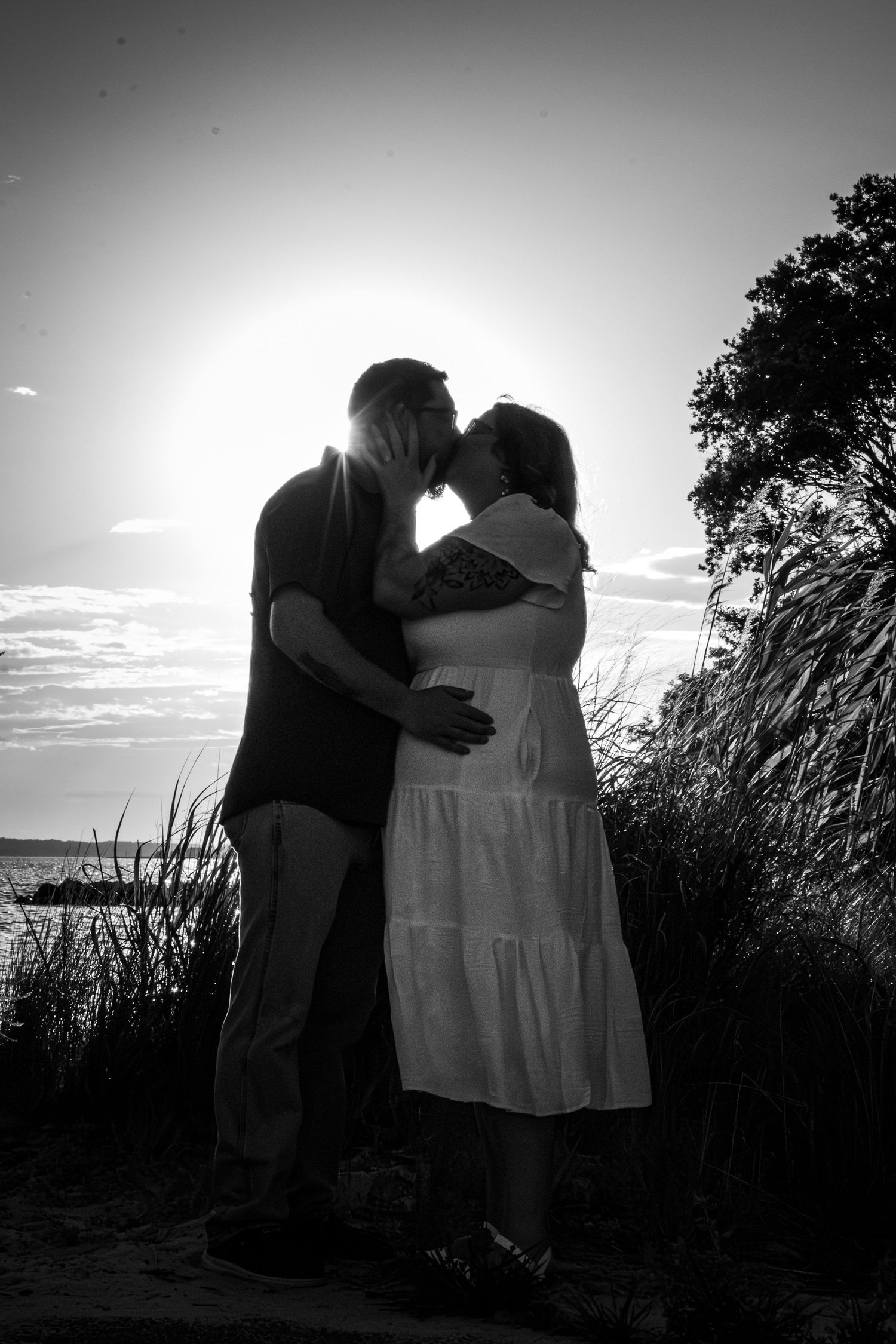Black and white silhouette of a couple kissing at sunset near the beach, surrounded by tall grass and trees.