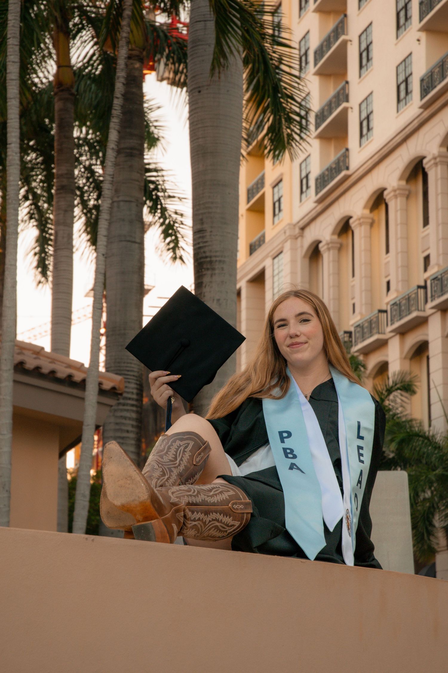 Young woman in graduation gown and cap holding mortarboard, wearing PBA sash, posing outdoors near palm trees.
