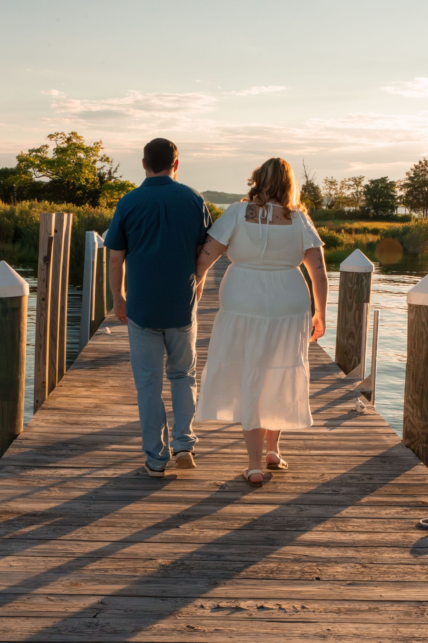 Couple holding hands walking on wooden dock at sunset, man in blue shirt and woman in white dress.