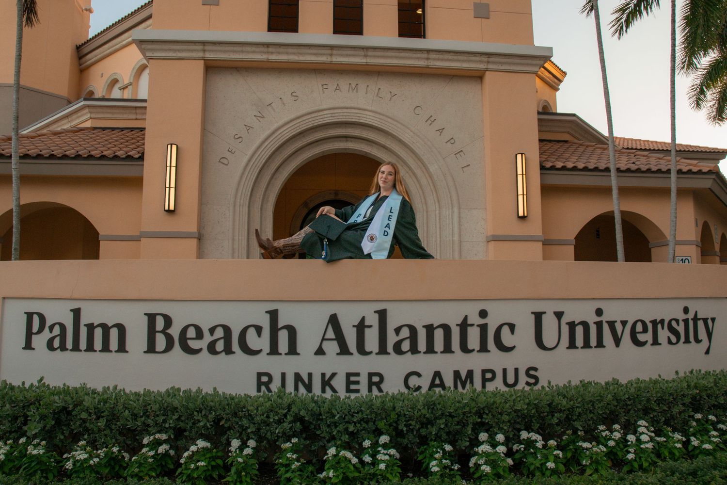 Graduate in cap and gown posing at Palm Beach Atlantic University Rinker Campus entrance sign.