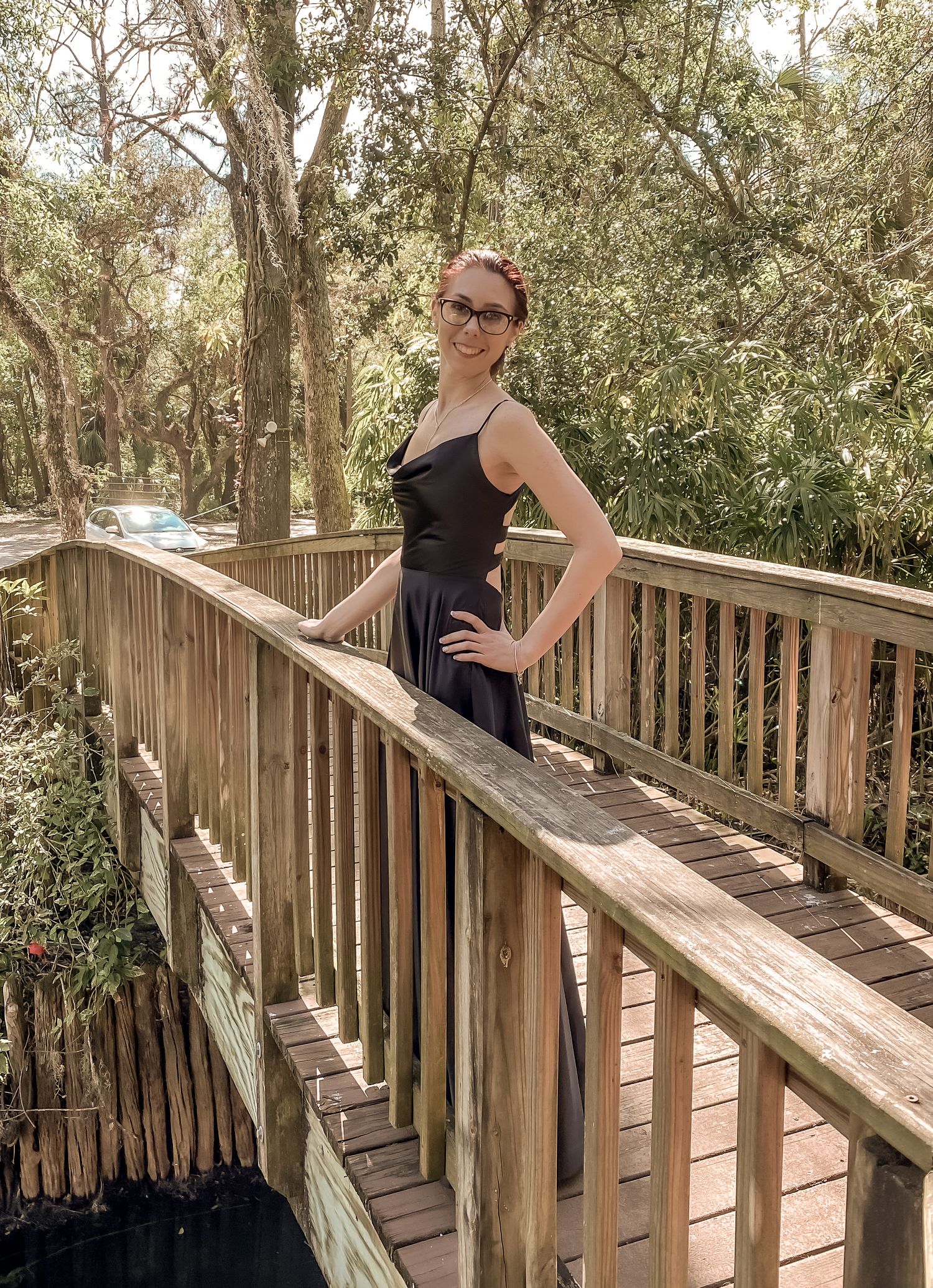 Woman in black dress posing on wooden boardwalk surrounded by trees and natural scenery.