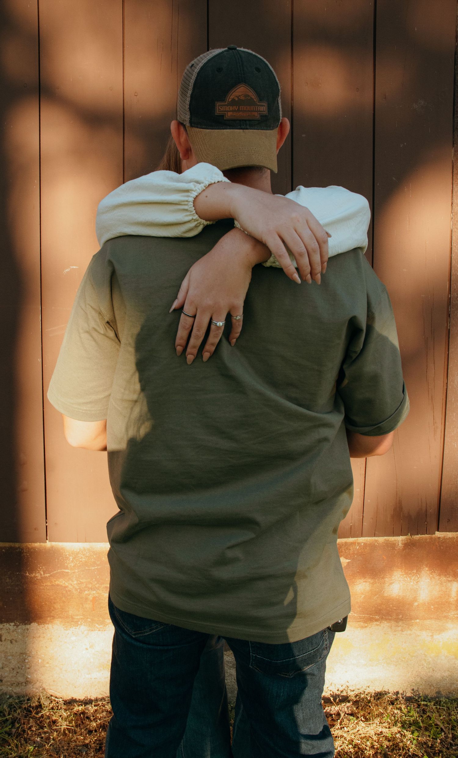 Two people sharing a warm embrace from behind, one wearing a green shirt and cap, the other with a ring on their finger.