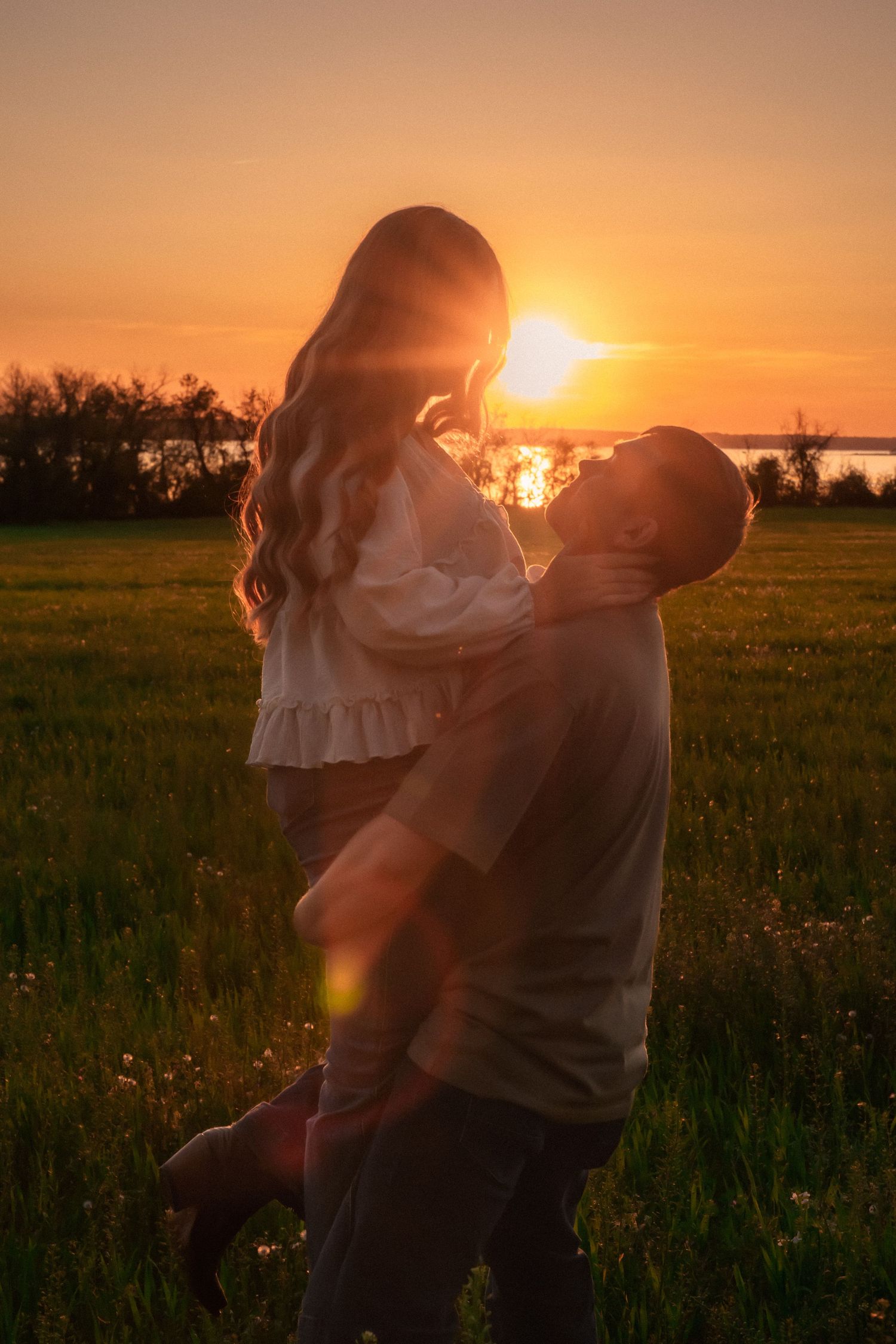 Romantic couple embracing in a sunlit field at golden sunset, silhouetted against vibrant orange sky.
