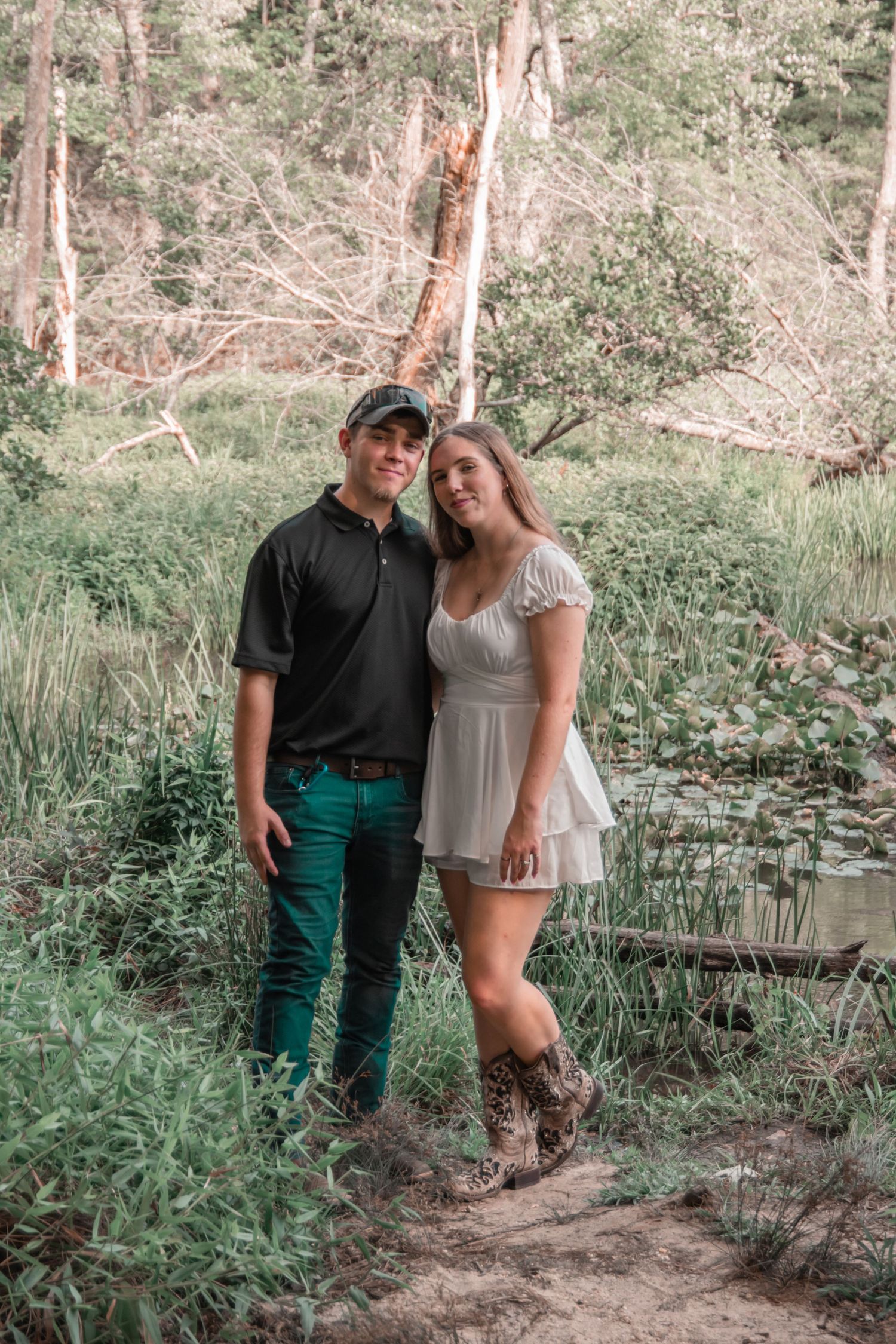 Couple posing together on nature trail with trees and pond in background.