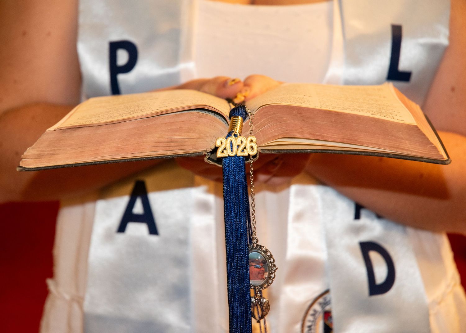 Student holding open book with blue tassel and 2026 graduation charm, wearing white graduation stole.