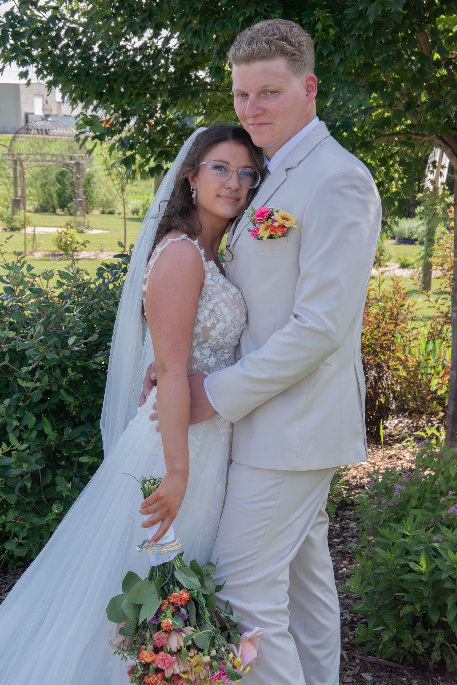 Bride and groom pose in garden, she holds colorful bouquet, he wears cream suit with pink boutonniere.