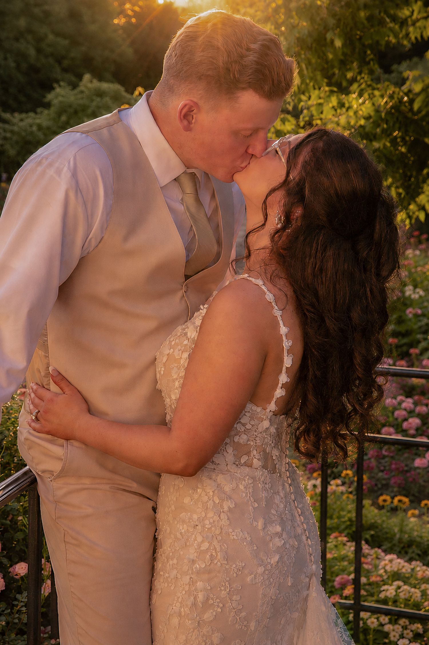Bride and groom share a romantic kiss at golden hour in a lush garden, bride in lace gown, groom in tan vest.