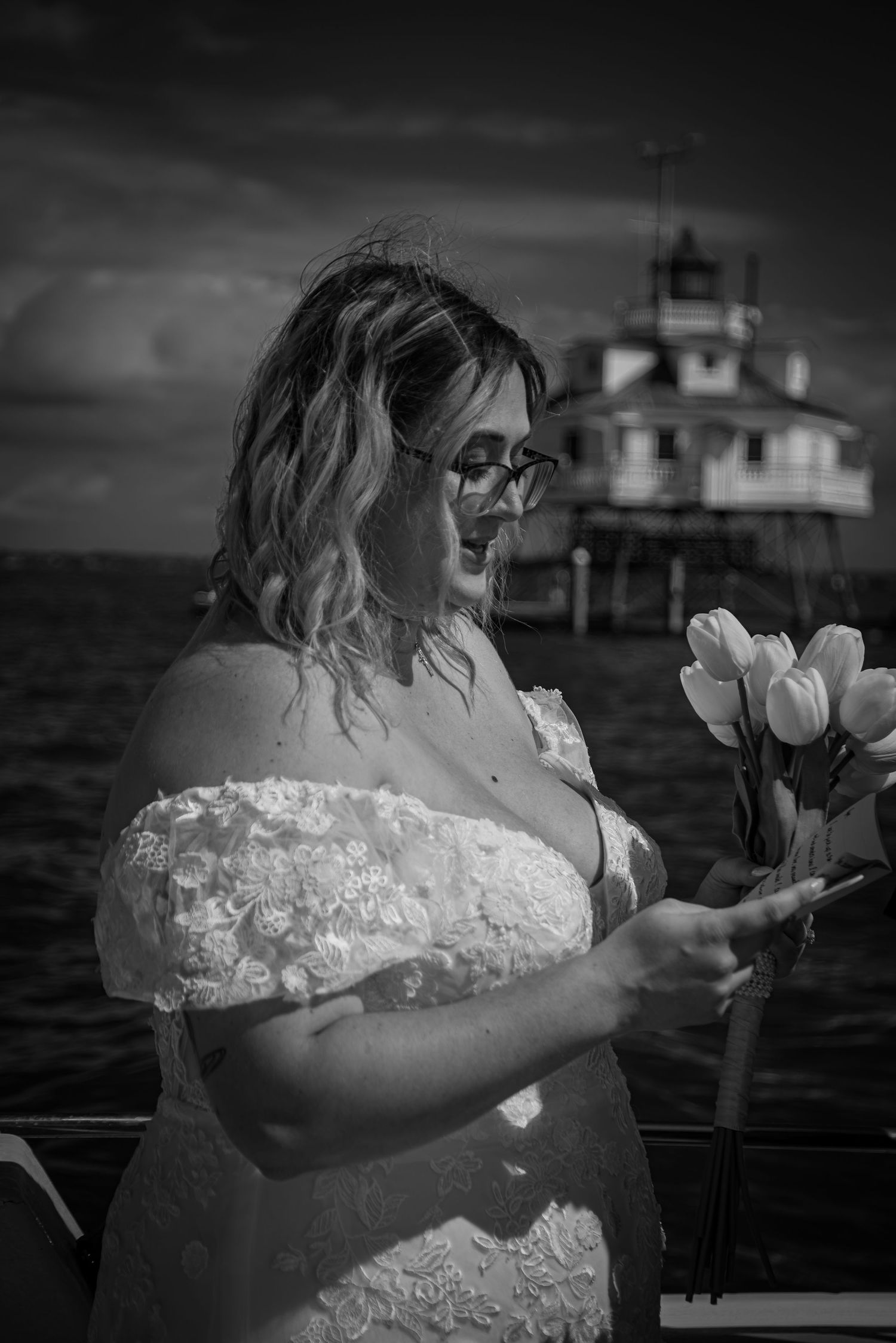 Bride in white off-shoulder lace gown holds tulips near a lighthouse on the water in black and white photo.