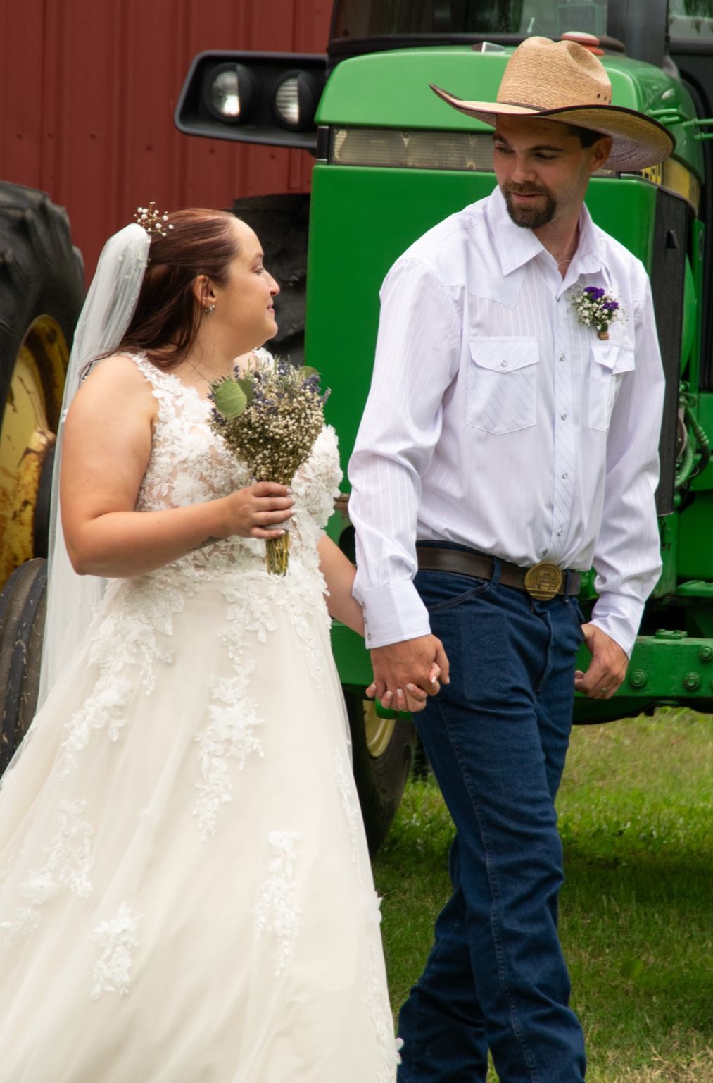 A couple in western wedding attire stands together outdoors with a green tractor visible in the background.