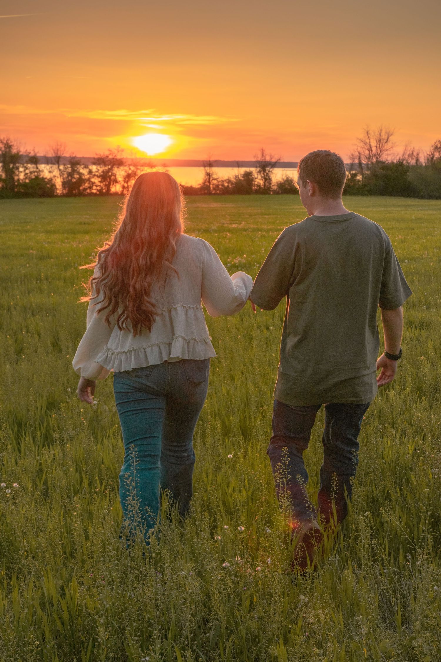 Couple holding hands walking through green field toward vibrant orange sunset near lake.