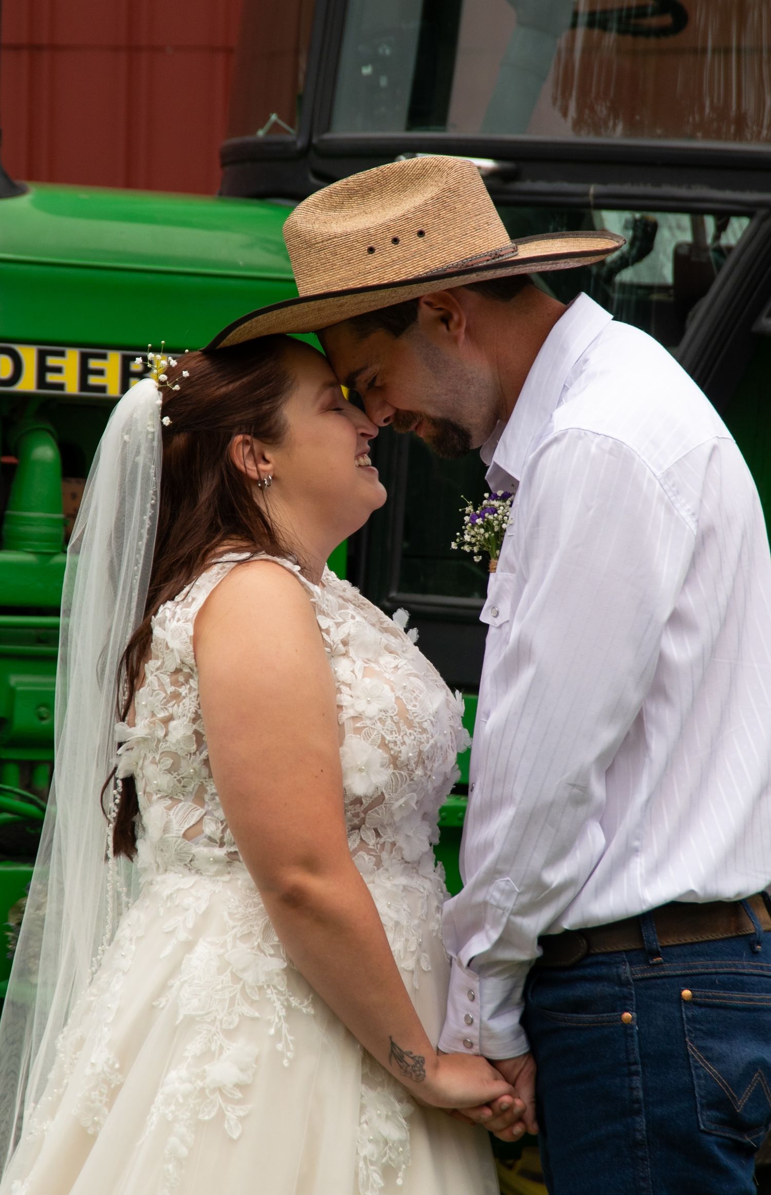 A tender wedding moment between a bride in a lace dress and groom in a cowboy hat with a John Deere tractor visible behind them.