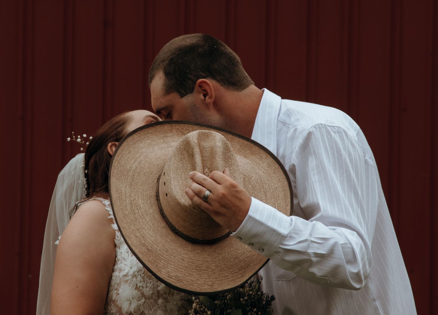 Intimate wedding moment captured against red barn backdrop with cowboy hat detail.