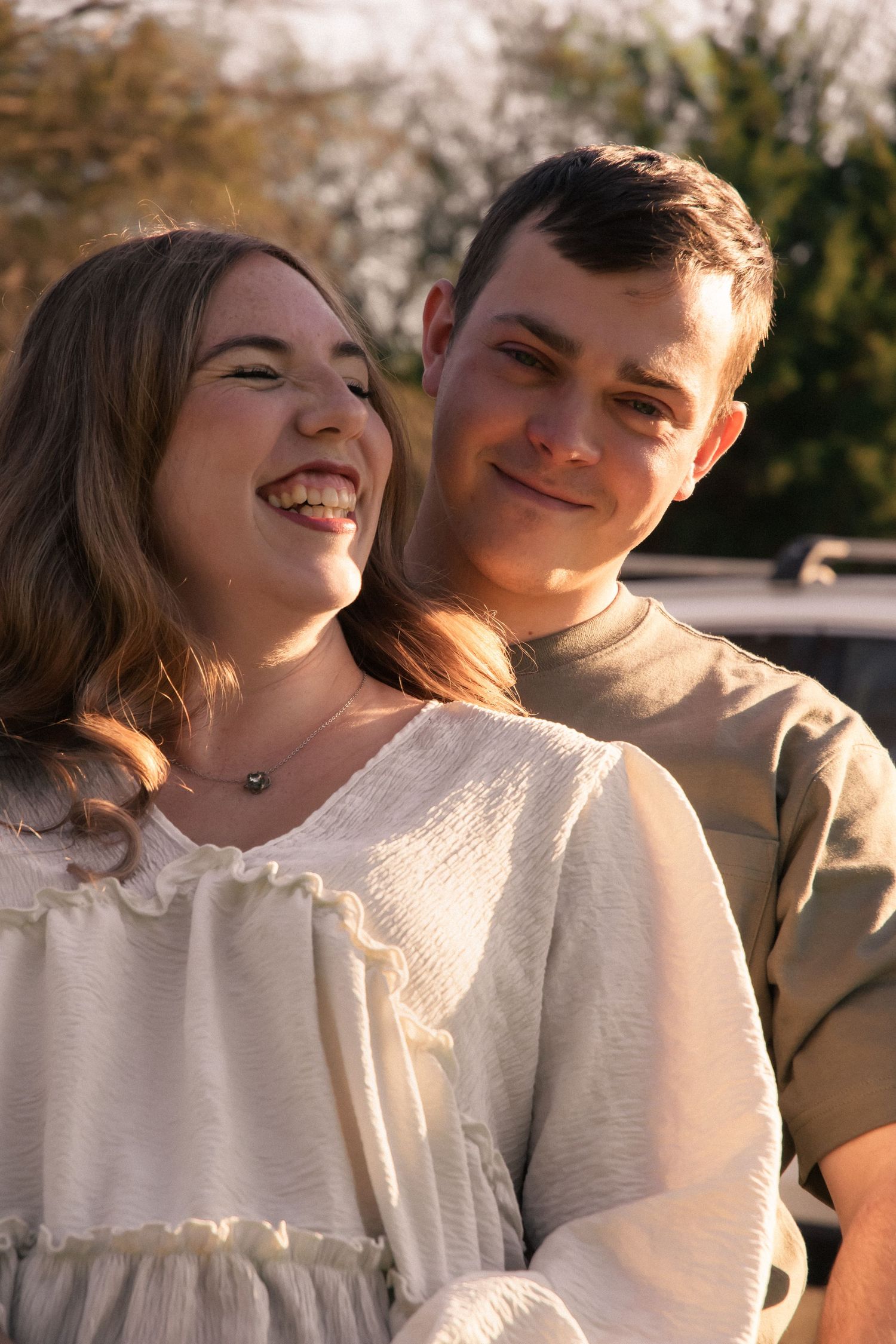 Young couple smiling and laughing together outdoors in golden hour sunlight, woman in white blouse and man in olive shirt.