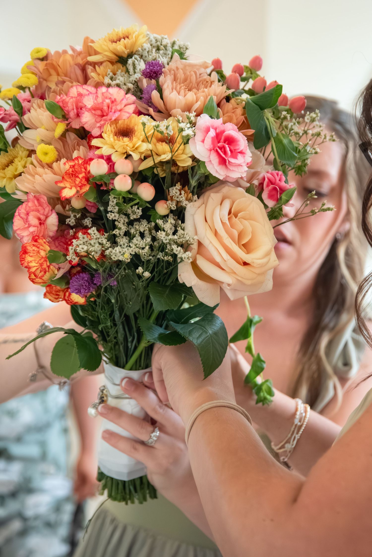 Woman holding a vibrant bridal bouquet with peach roses, pink carnations, and colorful wildflowers at a wedding.