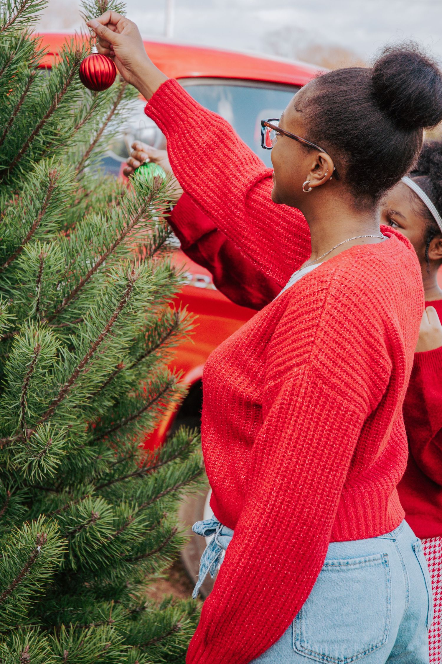 Person in red sweater hanging ornaments on evergreen Christmas tree branches.