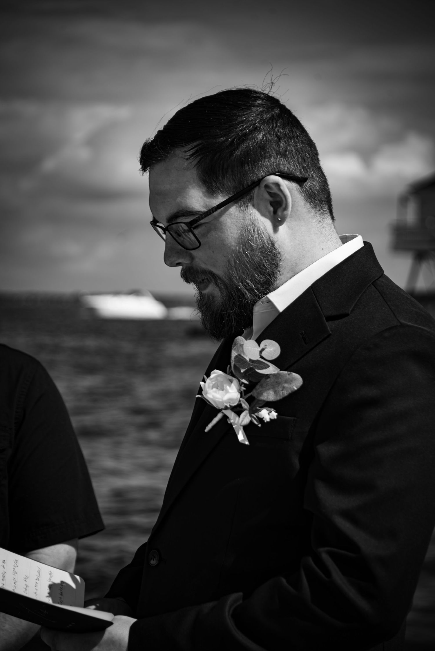 Black and white photo of a bearded groom in a suit with boutonniere reading vows outdoors near the ocean.