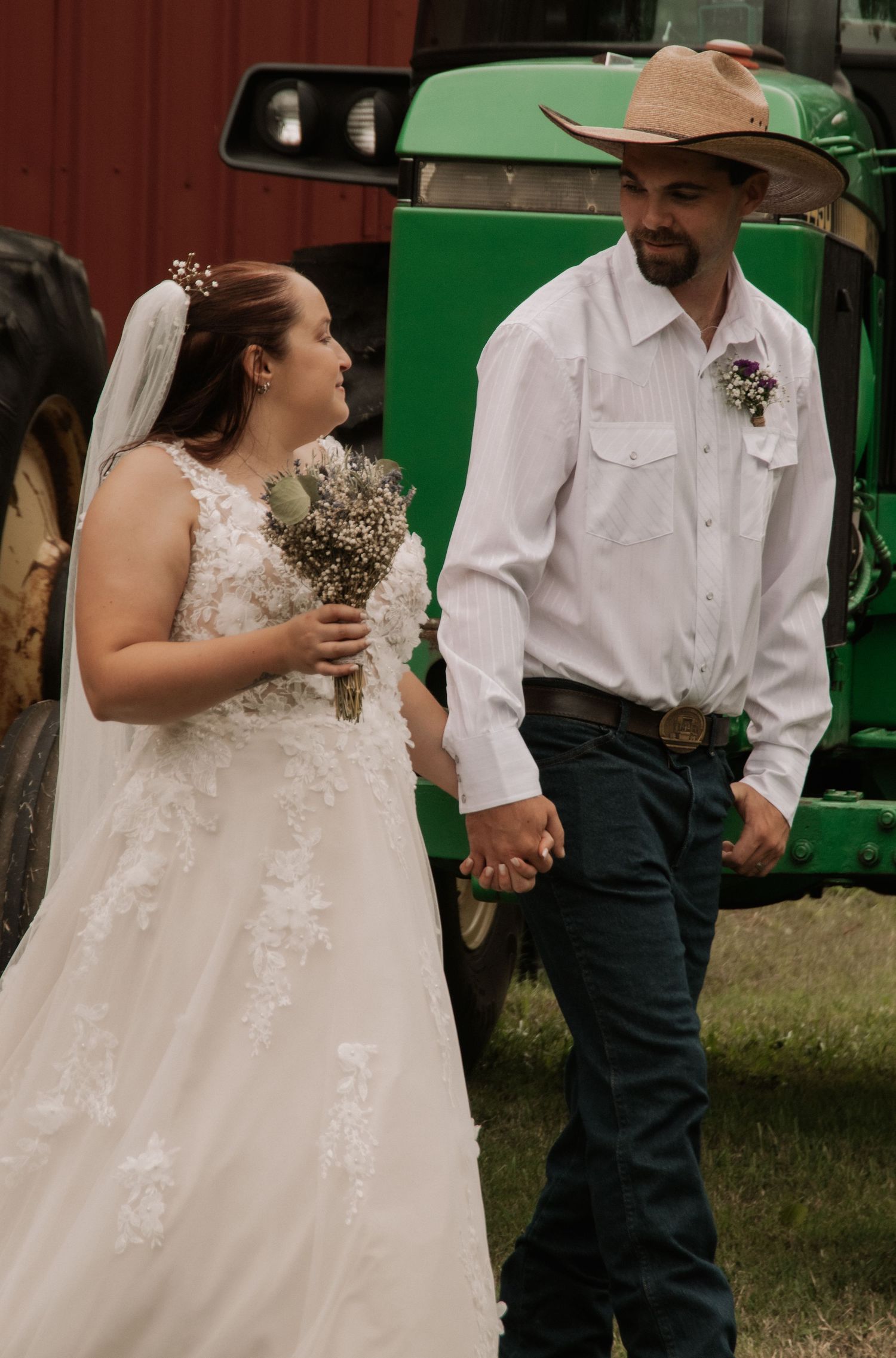 Country wedding couple in white dress and western attire near green tractors and red barn.