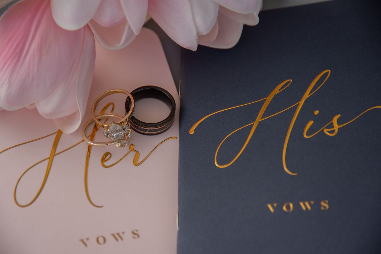 His and Her vows books with wedding rings, featuring gold script on pink and navy covers.