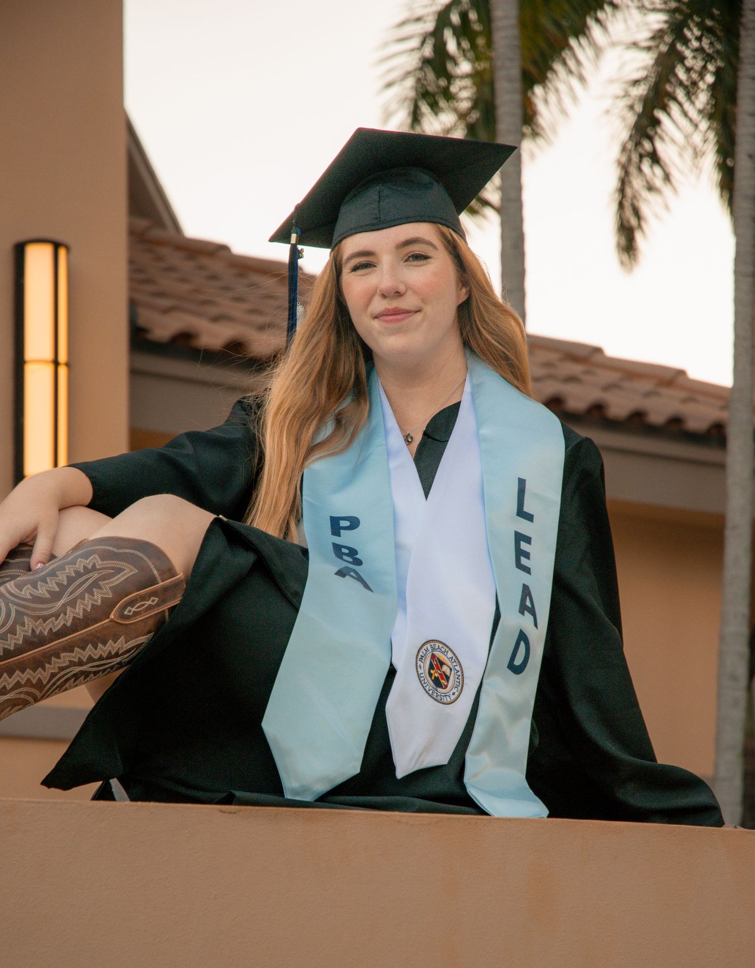 Smiling graduate in black cap and gown with LEAD stole poses outdoors near palm trees and terracotta building.