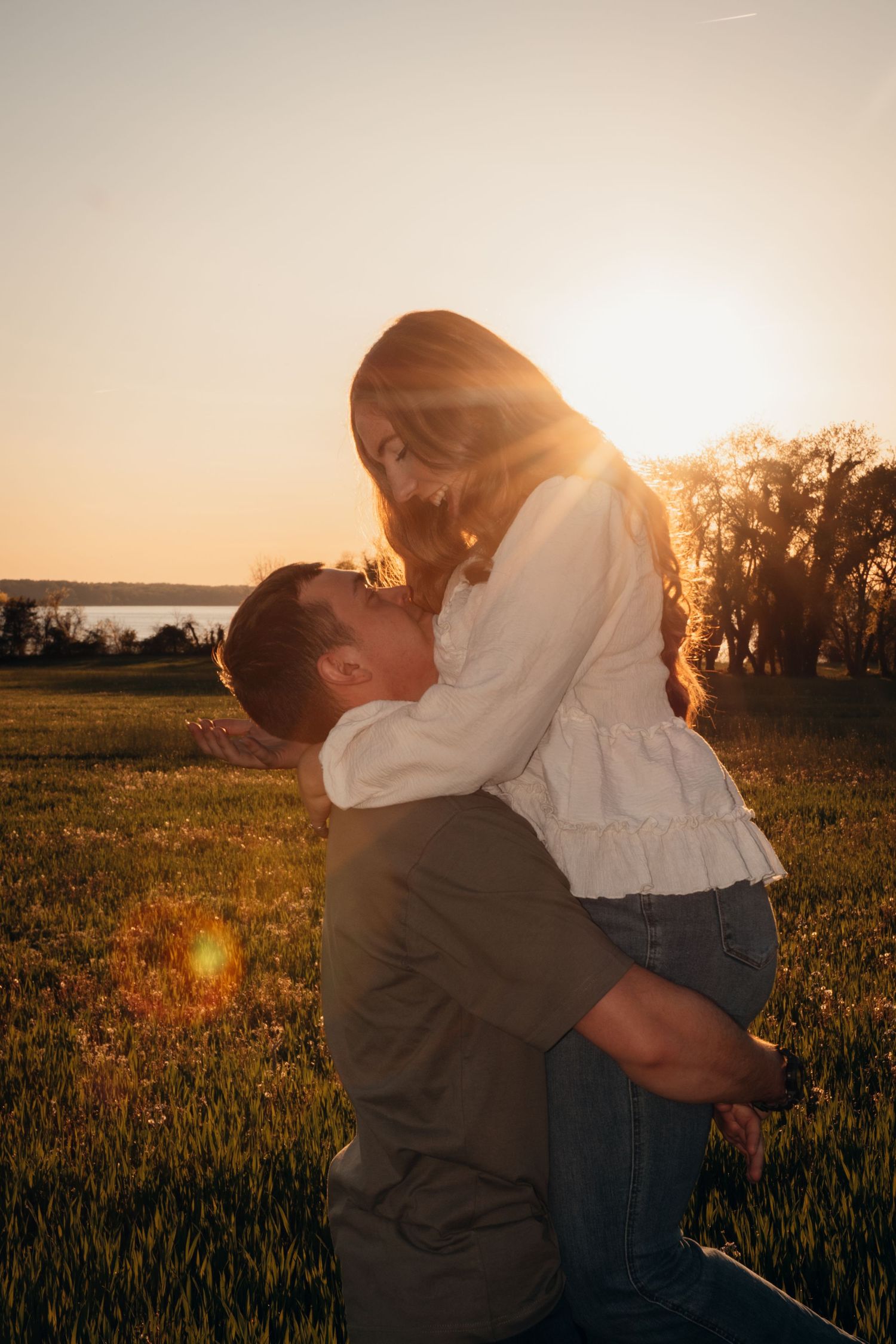 Couple sharing a romantic embrace at golden hour sunset in a green field near a lake.
