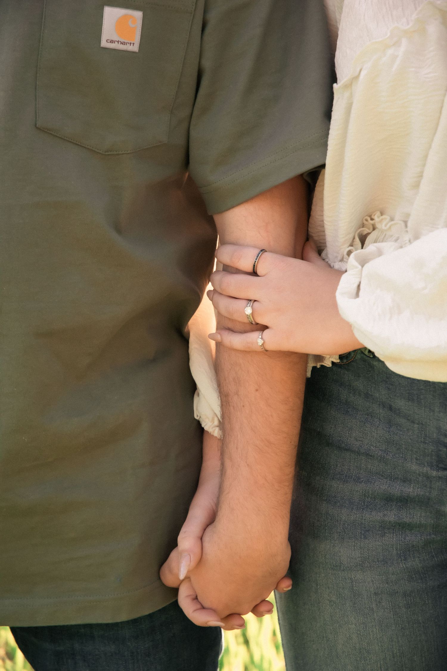 Couple holding hands showing engagement ring, woman wearing diamond ring, man in olive Carhartt shirt, romantic outdoor photo.