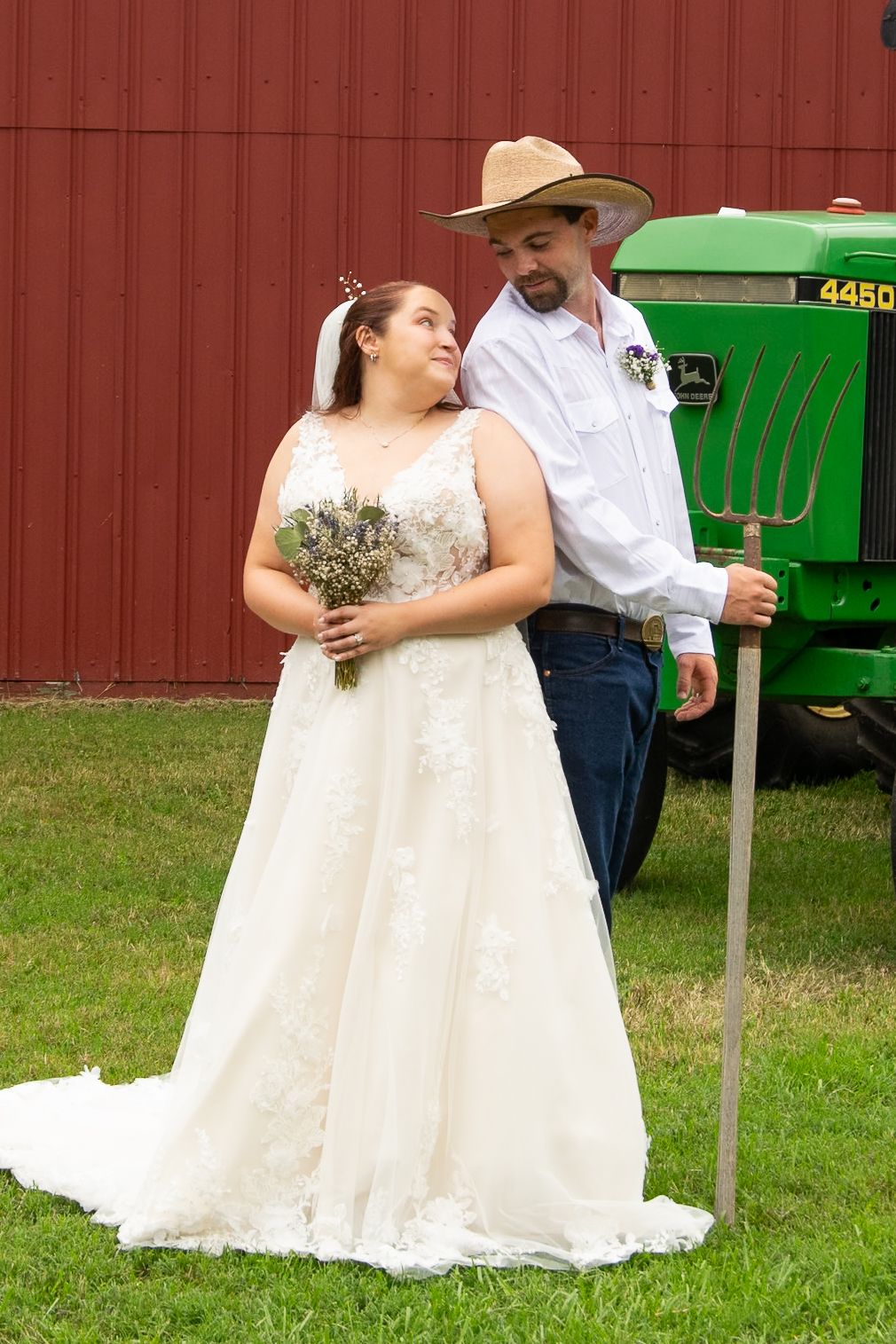 A couple poses for a wedding photo in front of a red barn wall and green John Deere tractor on a grassy lawn.