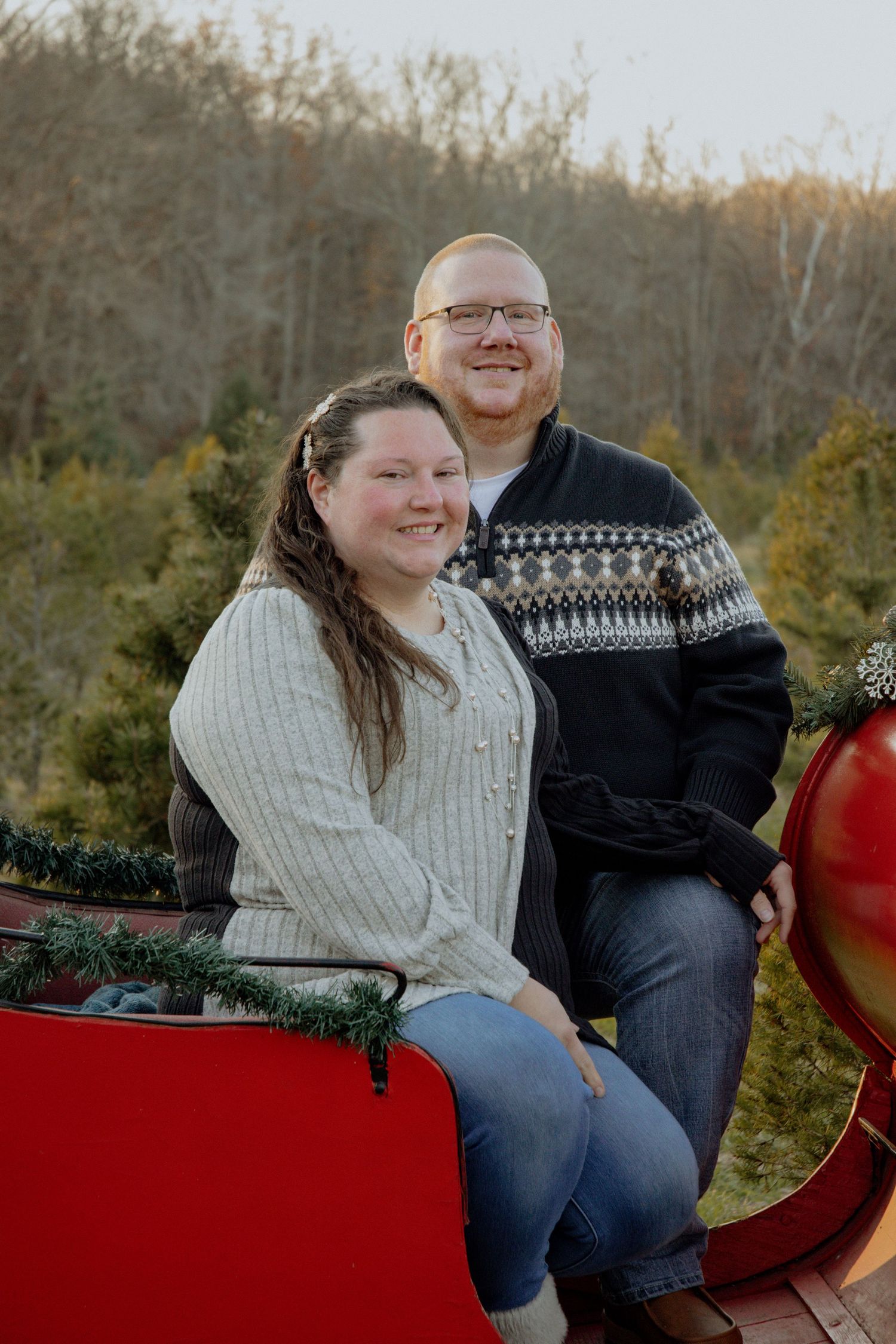 Happy couple sitting together in a red vintage sleigh surrounded by pine trees.