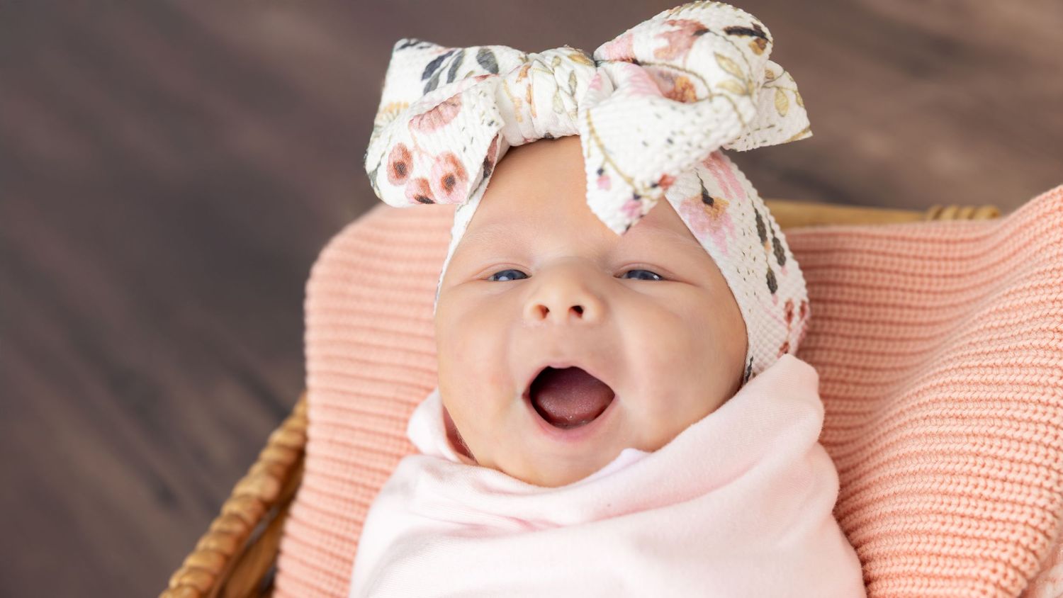 A baby wearing a white bow headband and peach-colored outfit smiles while laying on a textured surface.