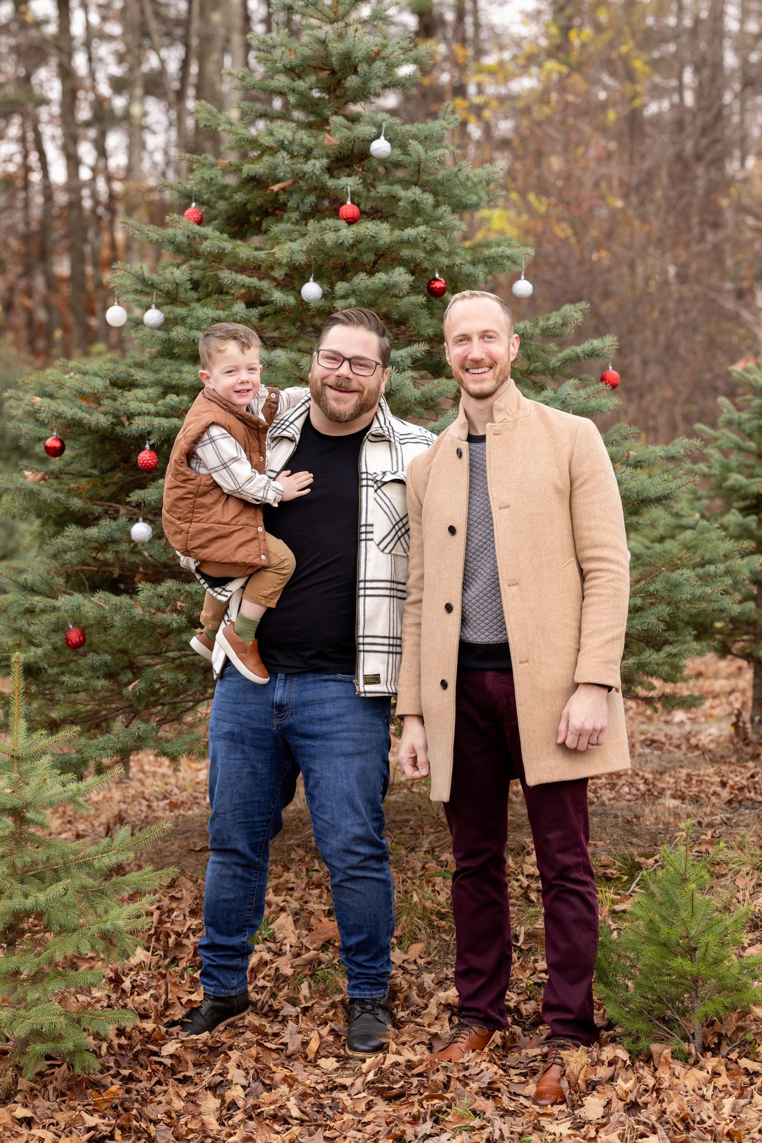 A group in coordinating outfits stands together among evergreen trees decorated with red ornaments for holiday photos.
