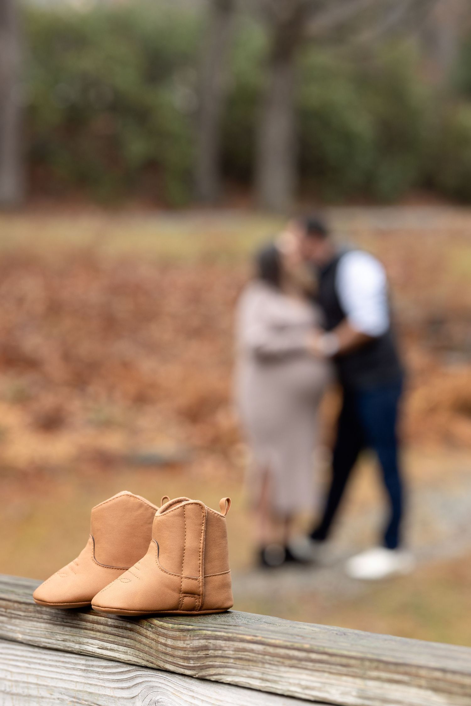 Tan leather baby booties sit on wooden deck boards with a blurred engagement photoshoot scene in the background.